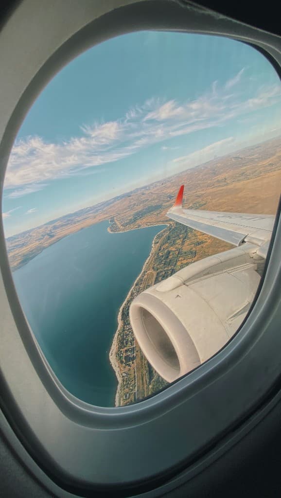 View from an airplane window of blue water and arid coastline, with the wing and engine in the foreground
