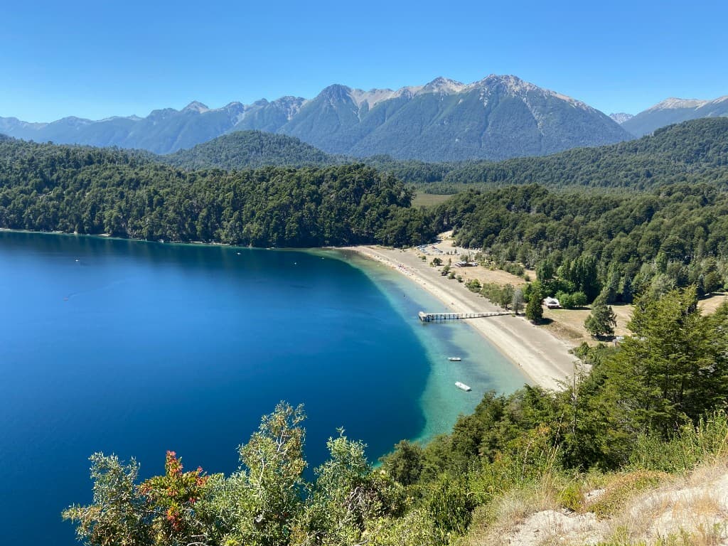 Elevated view of a turquoise lake and sandy beach bordered by dense forest and jagged mountains in Patagonia, Argentina